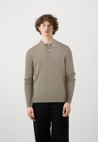 Polo beige a maniche lunghe con colletto, apertura a tre bottoni e orlo a coste. Indossato con pantaloni neri. Texture liscia.