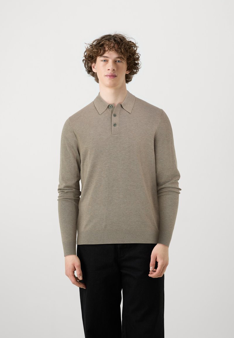 Polo beige a maniche lunghe con colletto, apertura a tre bottoni e orlo a coste. Indossato con pantaloni neri. Texture liscia.