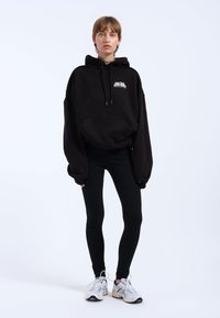 Jeune personne debout portant un sweat à capuche noir oversize avec texte, des leggings noirs et des baskets blanches sur un fond clair uni.