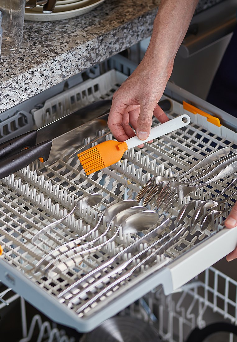 Geschirrspüler-Besteckkorb mit Edelstahlbesteck. Neben Gabeln und Löffeln wird ein oranger Silikonpinsel platziert. Strukturierte Kunststoffgestaltung.