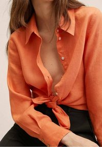 Femme portant une chemise orange déboutonnée nouée à la taille, associée à un pantalon noir taille haute, assise les cheveux détachés.