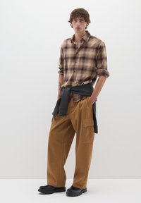 Camicia a quadri marrone con bottoni, felpa nera annodata in vita, pantaloni cargo beige e scarpe nere, con una vestibilità comoda e tasche.