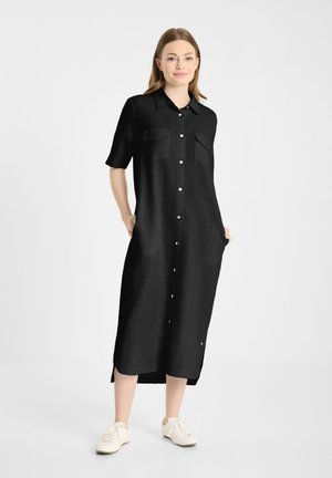 Vrouw staat, draagt een zwart midi overhemdjurk met knopen, korte mouwen en witte sneakers tegen een effen lichte achtergrond.
