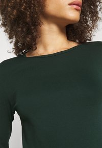 Mörkgrön långärmad topp med rundad halsringning, tillverkad av ett slätt tyg. Närbild på axeln och detaljerna vid halsringningen.