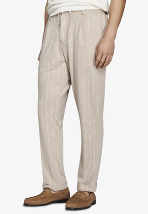 Uomo che indossa pantaloni beige a righe sottili con risvolti e mocassini di camoscio marroni, in piedi contro uno sfondo semplice.