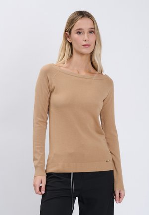 Frau mit langen blonden Haaren, die einen camel-farbenen Pullover mit Boot-Ausschnitt und schwarze Hose vor hellem, einfarbigem Hintergrund trägt.