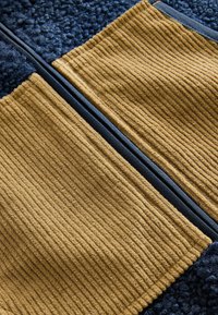Pannello in tessuto con materiale a coste senape, accentuato da un tessuto navy testurizzato. Un bordo navy liscio delimita la cucitura tra i materiali.