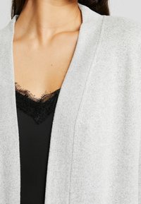 Lichtgrijze cardigan gemaakt van zachte stof, met een open voorkant en een gestructureerde zwarte kanten rand die onder een zwart shirt uitsteekt.