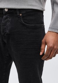 Κοντινό πλάνο ενός ατόμου που φοράει μαύρο τζιν και γκρι μακρυμάνικο πουκάμισο, με το χέρι χαλαρό δίπλα στο σώμα.