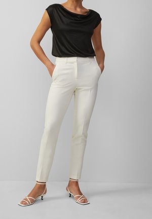 Femme portant un haut noir sans manches, un pantalon blanc slim taille haute, et des sandales blanches à brides avec un petit talon, debout les mains dans les poches.