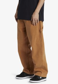 Pantalones holgados de color caramelo hechos de algodón pesado. Cuentan con bolsillos laterales y detalles de costura. Llevados con zapatillas negras.