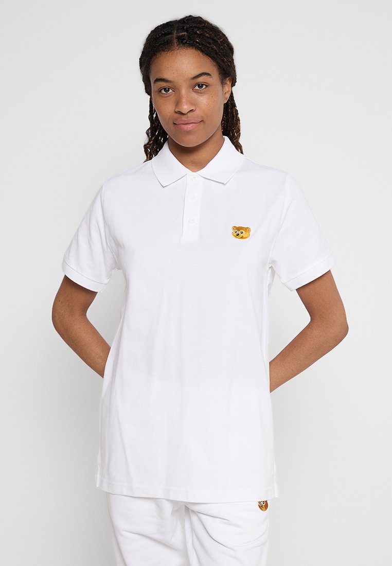 Camisa polo blanca de algodón, con cuello de tres botones y un pequeño parche bordado de un oso en el pecho. Mangas cortas y corte relajado.