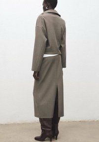 Khaki płaszcz wełniany z kołnierzem, opuszczonymi rękawami i rozcięciem z tyłu. Noszony na białej bluzce, w połączeniu z brązowymi dopasowanymi kozakami sięgającymi do kolan.