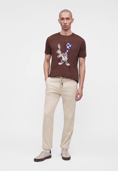Άντρας που φοράει καφέ t-shirt με πολύχρωμο καρτούν κουνέλι, μπεζ παντελόνι με κορδόνι και μπεζ αθλητικά παπούτσια με σκούρα σόλα.