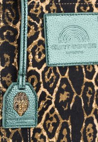 Tessuto con stampa leopardata e dettagli dorati; etichetta in pelle verde testurizzata con un charm decorativo dorato e un logo impresso, creando un contrasto mozzafiato.