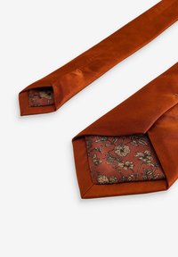 Satin slips i rustfarve med en tekstureret overflade. Den indre foring har et blomstermønster i guld og mørkerød på en ensfarvet baggrund.