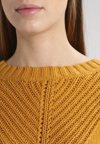 Senfgelbes Strickpullover mit strukturiertem Zickzack-Muster und geripptem Kragen. Nahaufnahme, die sich auf die Stoffdetails konzentriert.