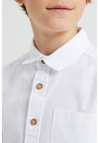 Bílá košile s límečkem a knoflíkovou přední částí, se dřevěnými knoflíky a kapsou na prsou. Tkanina vypadá lehce a má texturovaný povrch.