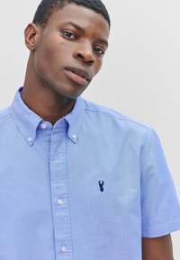 Chemise bleu clair à manches courtes avec col et petit logo homard violet sur la poitrine. Texture du tissu lisse.