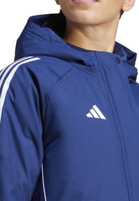 Dunkelblaue Jacke mit Kapuze, mit weißen Streifen an den Schultern und einem kleinen weißen Adidas-Logo auf der linken Brust. Glatte Textur.