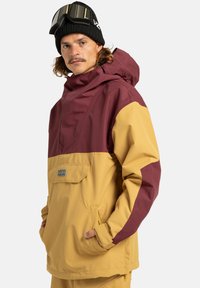 Farbblockierte Skijacke mit oberem Teil in Kastanienrot und unterem Teil in Senfgelb. Verfügt über eine Kapuze, eine Vordertasche und eine weiche, wasserabweisende Stoff.