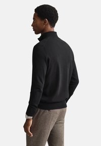 Crni pleteni pulover s ovratnikom na patentni zatvarač, s dugim rukavima i rebrastim rubom, uparen s smeđim hrapavim hlačama.