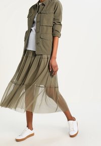 Chaqueta verde oliva con dos bolsillos delanteros, falda transparente en capas y zapatillas blancas con suela de goma. Top a rayas debajo de la chaqueta.