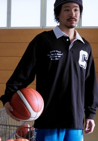 Camisa polo negra de manga larga con cuello blanco, que presenta detalles de logotipo y texto. Hombre sostiene un baloncesto, vistiendo pantalones cortos azules. Ambiente de gimnasio cerrado.