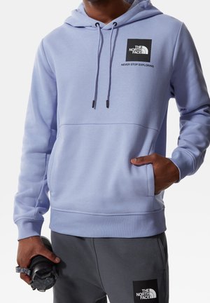 Person iført en lyse lilla hættetrøje og mørkegrå bukser med The North Face-logoer, der holder en sort vandflaske i den ene hånd.