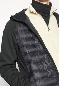 Svart långärmad jacka med en kviltad marinblå panel, lagd över en krämfärgad zip-up tröja med texturerad yta.