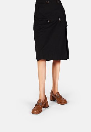 Mocassini in pelle marrone con tacco a blocco, punta quadrata e accentuazione metallica, abbinati a una gonna nera doppiopetto. Design con suola piatta.