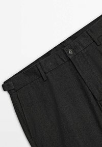 Gros plan sur la taille d’un pantalon habillé gris foncé avec fermeture par bouton, passants pour ceinture et motif subtil en chevrons sur fond blanc.