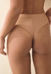 Beige naadloze thong ondergoed, met een hoge uitsnijding en een gladde textuur, die de achterkant en heupen toont met subtiele striae.