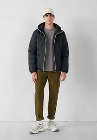 Uomo che indossa un cappello beige, giacca a vento nera, maglietta grigia, pantaloni oliva arrotolati, calze bianche e sneakers bianche, in piedi di fronte a una parete semplice.