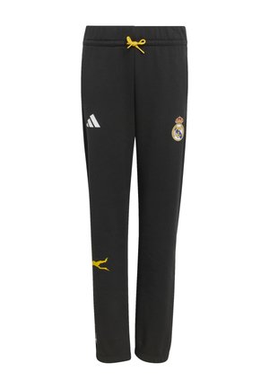 Dwoje dzieci stoi obok siebie, ubrane w czarne dresy Real Madryt z żółtymi i białymi wzorami graficznymi oraz butami marki Adidas.