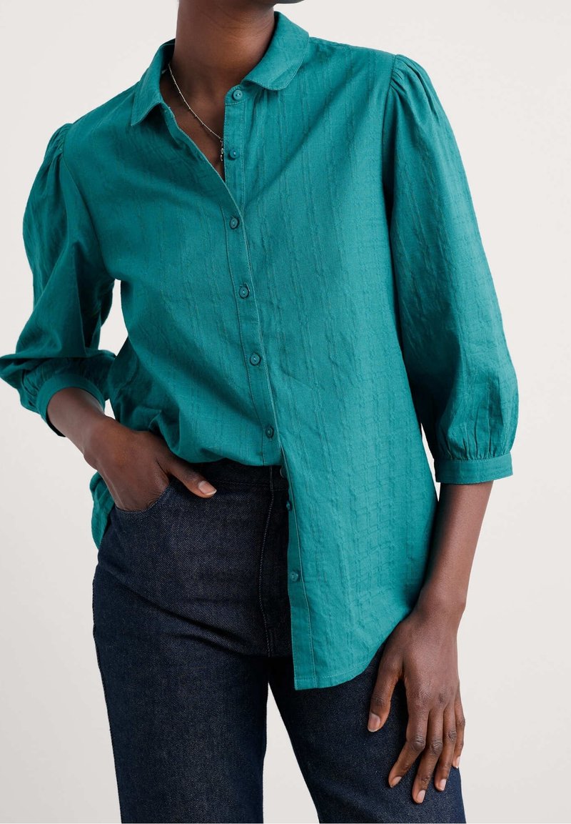 Personne portant une chemise boutonnée texturée bleu sarcelle avec des manches trois-quarts bouffantes et un jean bleu foncé, main dans la poche et pose détendue.