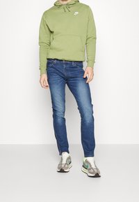 Grön hoodie med framficka och snöre. Mörkblå jeans. Grå, gröna och vita sneakers med texturerad sula och flärdfulla färgaccenter.