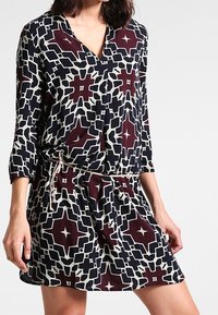 Tištěné šaty s výstřihem do V, tříčtvrtečními rukávy a páskem na stažení v pase. Barvy zahrnují námořnickou modrou, krémovou a bordó geometrické vzory.