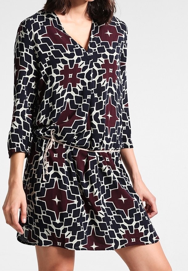 Tištěné šaty s výstřihem do V, tříčtvrtečními rukávy a páskem na stažení v pase. Barvy zahrnují námořnickou modrou, krémovou a bordó geometrické vzory.