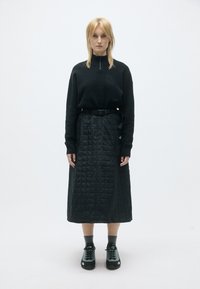 Top de fleece preto com fecho, adornado com um logótipo, combinado com uma saia midi preta acolchoada e ténis pretos com toques de verde claro.