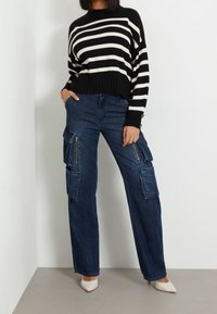 Maglione a righe bianche e nere con orlo a coste abbinato a pantaloni cargo larghi di colore blu scuro, dotati di molteplici tasche e cerniere.