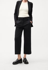 Sort skræddersyet blazer over en beige top, kombineret med sort højtaljede bukser og chunky sorte loafers. Glat stof, lige ben-silhuet.