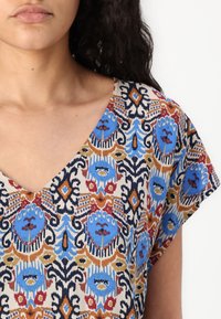 V-neck blouse featuring a patterned fabric with blue, red, and gold accents on a cream background; short sleeves and soft texture.