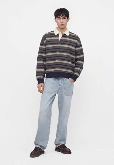 Homme portant un polo à manches longues rayé, un jean bleu clair ample et des chaussures marron foncé, debout devant un fond blanc uni.