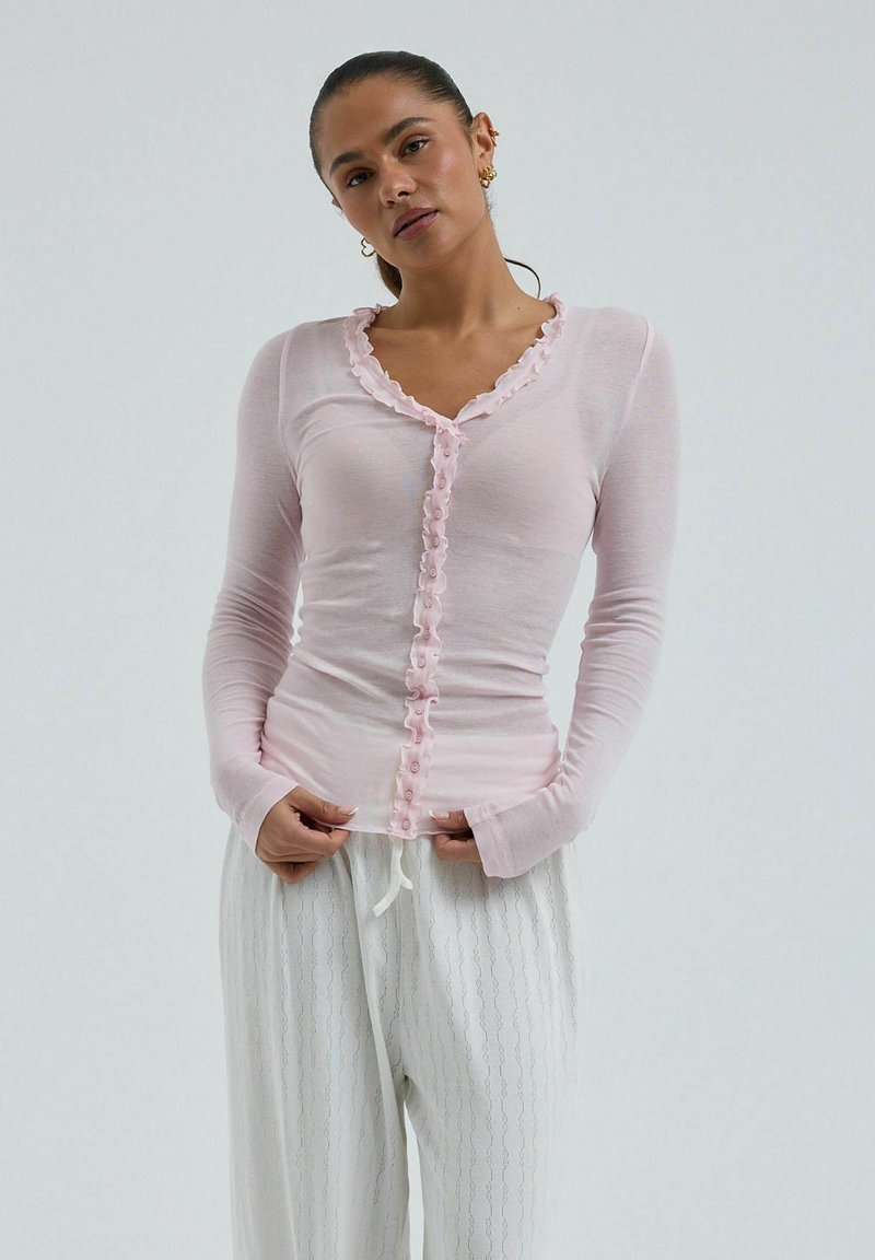 Femme portant un haut boutonné à manches longues rose clair avec des bords froncés et un pantalon blanc ample à motifs sur un fond uni.