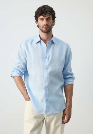 Hombre con cabello rizado y oscuro, vestido con una camisa de manga larga azul claro con las mangas remangadas y pantalones color crema, de pie frente a un fondo liso.