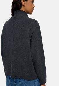 Kobieta w ciemnoszarej swetrze z wysokim kołnierzem i długimi rękawami, w niebieskich dżinsach, pokazana z tyłu i z boku na tle białym.