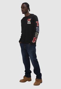 Mann mit geflochtenem Haar, der ein schwarzes Langarmshirt mit buntem Schriftzug auf dem Ärmel, blaue Jeans und beige Stiefel trägt und mit den Händen in den Taschen steht.