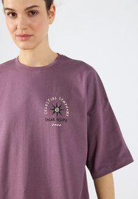 Violetti ylisuuri t-paita, joka on valmistettu pehmeästä kankaasta ja jossa on graafinen kuvasto, jossa on teksti "Taivaallinen Synafooni Auringonpimennys" sekä tähden kuvio.