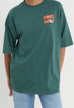 Personne portant un t-shirt oversize vert foncé avec "MORE AMOR" et des motifs floraux orange et violet sur la partie supérieure gauche de la poitrine.